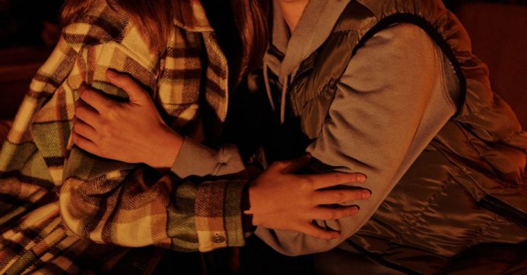 Free Camping - Positive female friends wearing warm clothes hugging and looking at camera while sitting near campfire in nature at evening time