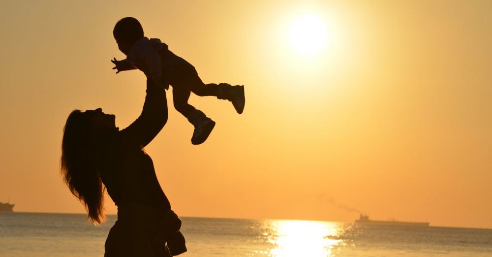 Family Beaches - Silhouette Photo of a Mother Carrying Her Baby at Beach during Golden Hour